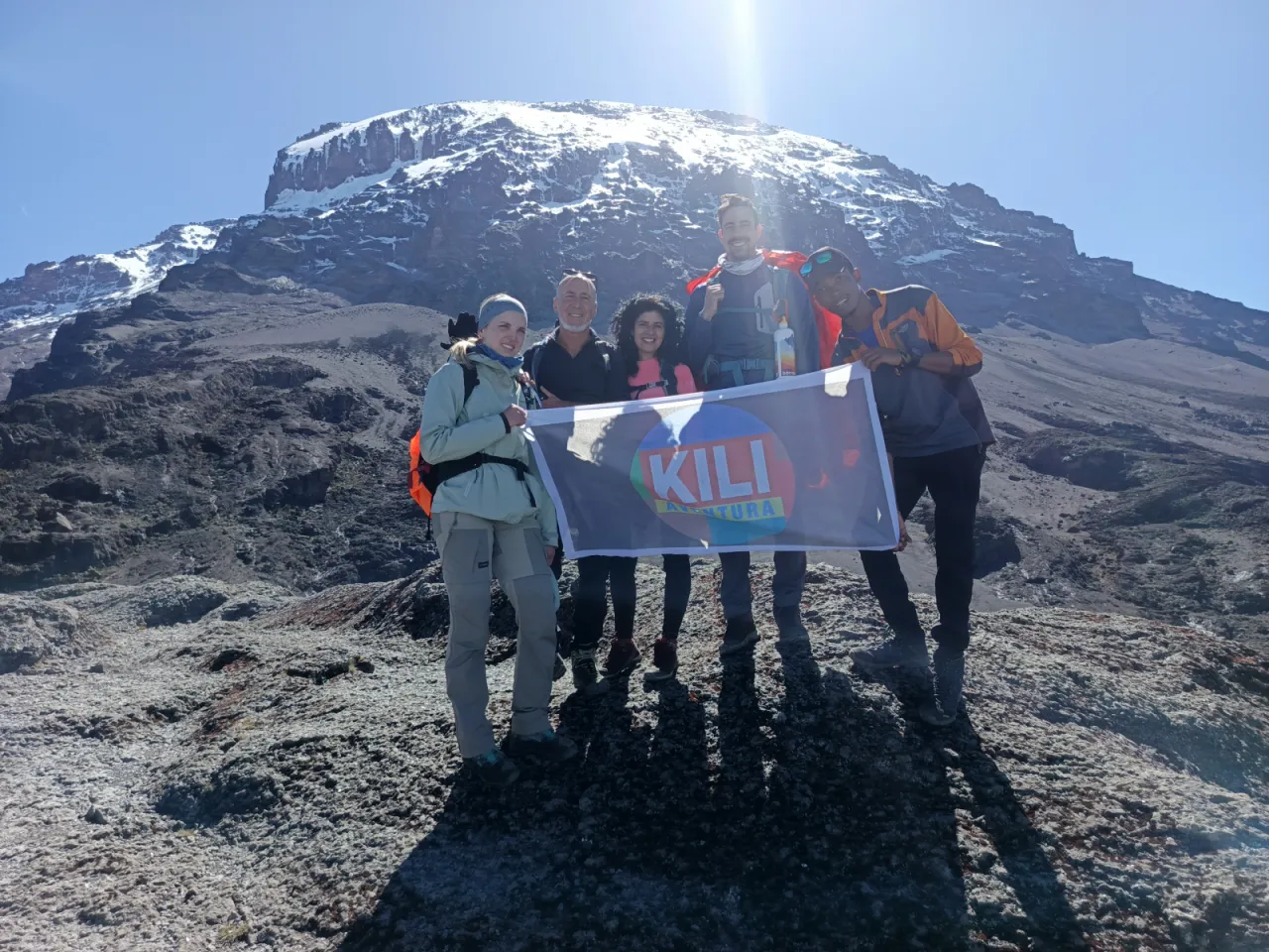 Five climbers holding the Kili Aventura banner on a rocky slope beneath Kibo's glaciers, with the sun behind the summit
