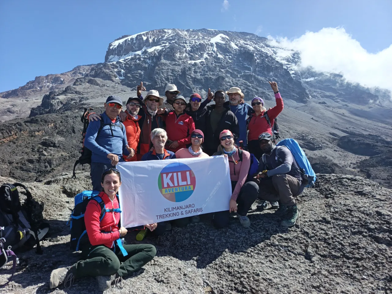 A smiling group of climbers and guides with the Kili Aventura banner on rocky slopes beneath Kibo's snow-capped summit