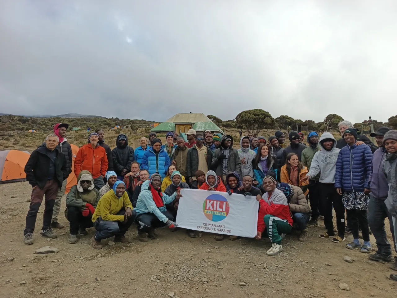 A large group of climbers and Kili Aventura crew gathered behind the company banner in a moorland camp