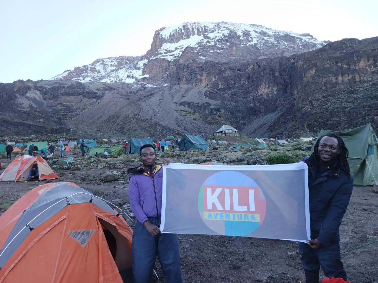 Two Kili Aventura crew members holding the company banner in a tented camp below the snow-streaked cliffs of Kibo