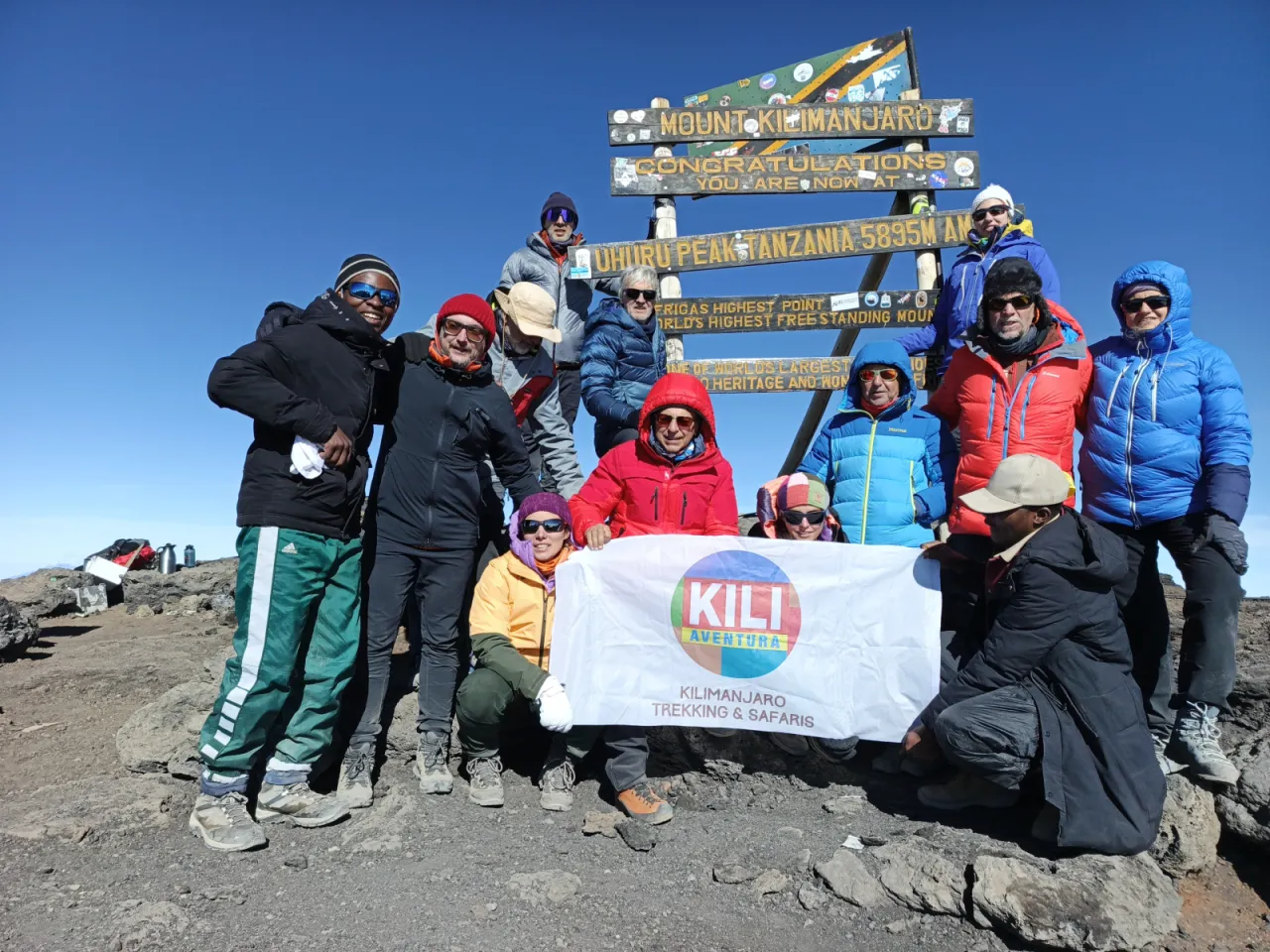 A group of climbers and guides with the Kili Aventura banner at the Uhuru Peak sign on the crater rim