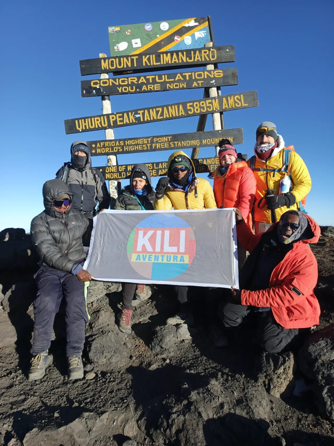 Climbers in down jackets with the Kili Aventura banner at the Uhuru Peak summit sign, 5,895 m, under a clear blue sky